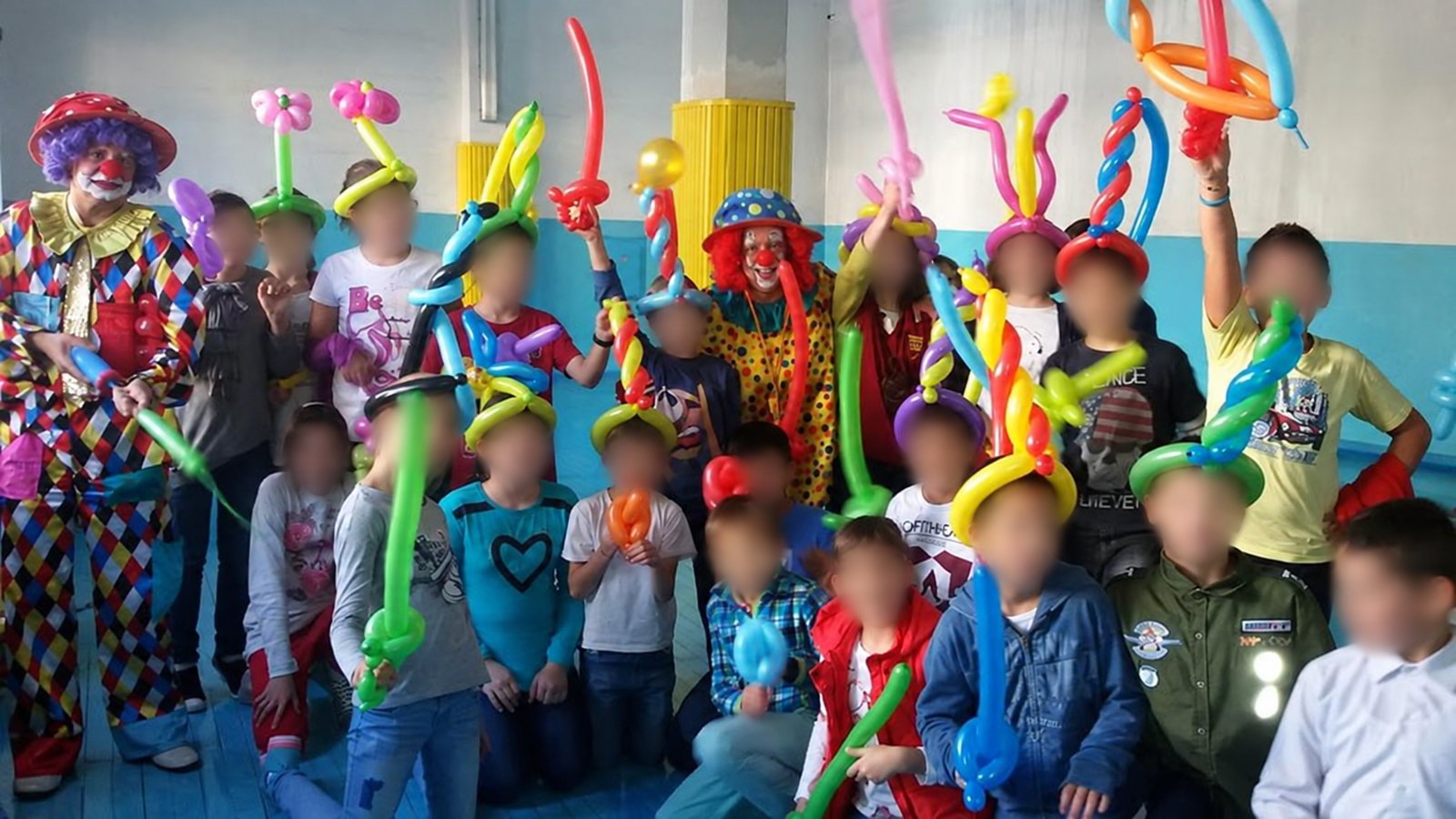Група деца държат балонни мечове, цветя и шапки, а двамата клоуни са в средата; лицата на децата са замъглени.